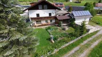 Luftaufnahme eines Hauses mit gepflegtem Garten, umgeben von grüner Landschaft und Bergen im Hintergrund.
