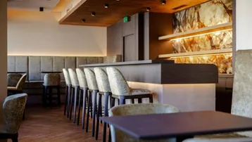 Moderne Bar mit dunklem Holz, gepolsterten Hockern und beleuchteter Steinwand, die eine warme Atmosphäre schafft.