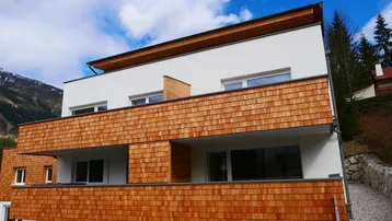 Modernes Mehrfamilienhaus mit Holzschindelfassade und Balkonen in alpiner Umgebung.