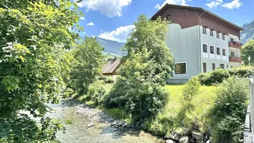 Gebäude am Flussufer mit üppiger Vegetation und Bergen im Hintergrund unter blauem Himmel.