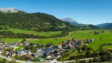 Malerische Landschaftsaufnahme mit einem Dorf, grünen Hügeln und Bergen im Hintergrund unter blauem Himmel.