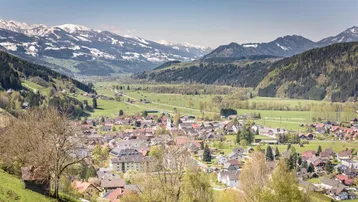 Malerische Ansicht eines Dorfes im Tal, umgeben von grünen Wiesen und schneebedeckten Bergen.