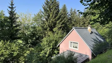 Kleines Haus mit rosafarbener Fassade und Satteldach, umgeben von üppigem Grün und hohen Bäumen.