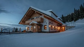 Alpenhaus bei Dämmerung mit beleuchteten Fenstern und schneebedeckter Umgebung.
