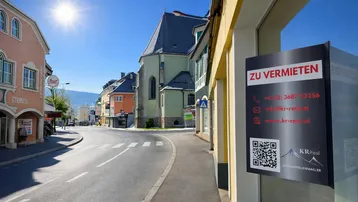 Sonnenbeschienene Straße in einer malerischen Stadt mit traditionellen Gebäuden und einem 'Zu Vermieten'-Schild im Schaufenster, im Hintergrund Berge.