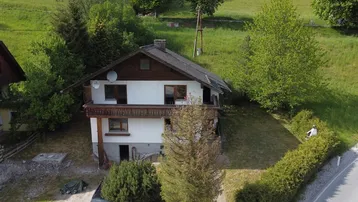 Ländliches Haus mit Balkon und Satteldach, umgeben von grünen Hügeln und Bäumen.