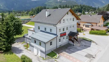 Luftaufnahme des Mehrfamilienhauses mit umliegender Landschaft und Bergen im Hintergrund.