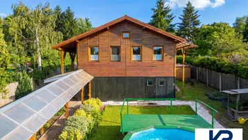 Holzhaus mit Garten, Pool und Solaranlage unter blauem Himmel, umgeben von viel Grün.