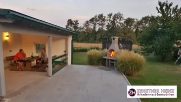 Gemütlicher überdachter Sitzbereich im Freien mit Holztisch und beleuchtetem Grill am Abend.