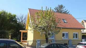 Gelbes Haus mit rotem Ziegeldach, blühendem Baum und einer Holzpergola an der Seite.