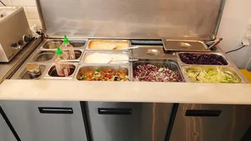 Edelstahl-Salatbar mit verschiedenen frischen Zutaten und Saucen für die Zubereitung von Speisen.