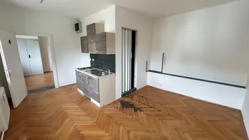 Helles Zimmer mit kleiner Küchenzeile, Fischgrätparkett und Zugang zu einem weiteren Raum.