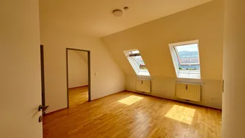 Helles Zimmer im Dachgeschoss mit Holzboden, Dachfenstern und Blick auf die Umgebung.