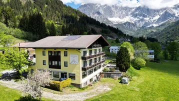 Luftaufnahme eines gelben Gebäudes mit Balkonen und Solarpaneelen, umgeben von grünen Wiesen und Bergen.