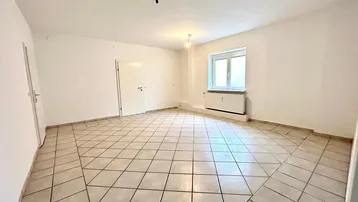 Heller, leerer Raum mit Fliesenboden, weißen Wänden und einem Fenster mit Heizkörper.