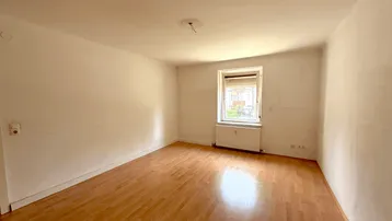 Helles, leeres Zimmer mit Laminatboden und einem Fenster mit Rollladen.