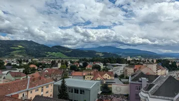 Panoramablick über die Stadt mit roten Dächern und majestätischen Bergen im Hintergrund unter bewölktem Himmel.