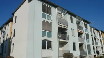 Mehrfamilienhaus mit mehreren Balkonen und heller Fassade unter blauem Himmel.