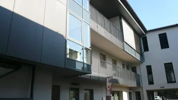 Moderne Gebäudefassade mit großen Fenstern, Balkonen und einem überdachten Eingangsbereich in hellem und dunklem Grau.