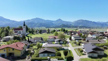Weitläufiger Blick über eine ländliche Siedlung mit Kirche und majestätischer Bergkulisse im Hintergrund.