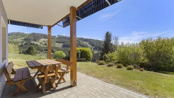 Überdachte Terrasse mit Holztisch und Bänken, umgeben von Natur und Bergblick.