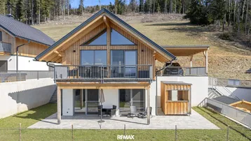 Modernes Holzhaus mit großem Balkon und Terrasse, umgeben von grüner Landschaft und Hügeln.