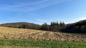 Blick über ein gepflügtes Feld mit grünen Akzenten und bewaldeten Hügeln im Hintergrund unter blauem Himmel.