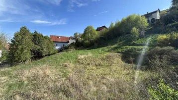 Grünes Hanggrundstück mit Blick auf die umliegende Bebauung und bewaldete Hügel unter blauem Himmel.