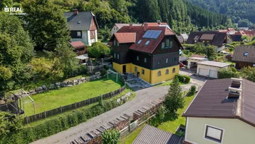 Luftaufnahme eines charmanten Hauses mit gelber Fassade, Holzelementen und Photovoltaikanlage, umgeben von einem gepflegten Garten und grüner Landschaft.