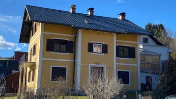 Gelbes Einfamilienhaus mit dunklem Satteldach und traditionellen Fensterläden unter blauem Himmel.