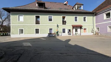 Traditionelles Mehrfamilienhaus mit hellgrüner Fassade, mehreren Fenstern und kleinen Balkonen an einem sonnigen Tag.