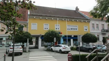 Gelbes Gebäude mit traditioneller Architektur, weißen Fensterläden und einem Durchgang zur Straße. Mehrere Autos sind vor dem Gebäude geparkt.