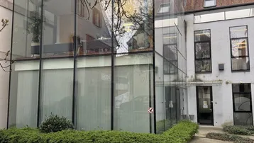 Weitwinkelansicht der modernen Glasfassade eines Geschäftsgebäudes mit umliegender Bepflanzung und einem Gehweg.