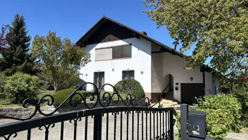 Das Einfamilienhaus hinter einem schmiedeeisernen Tor mit gepflasterter Auffahrt und grüner Umgebung.