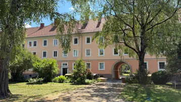 Mehrstöckiges Wohngebäude mit hellrosa Fassade, grüner Umgebung und einem Torbogen.