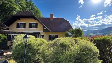 Traditionelles Haus mit gelber Fassade, rotem Dach und üppiger grüner Umgebung unter strahlendem Sonnenschein.