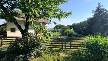 Hausansicht durch einen Baum, mit Holzzaun und weitem Blick auf die grüne Hügellandschaft.