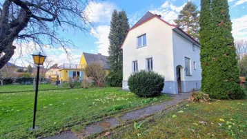 Weißes Einfamilienhaus mit großem Garten und altem Baumbestand unter blauem Himmel.