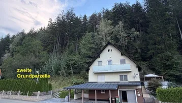 Ansicht des Hauses mit Carport und bewaldetem Hang im Hintergrund, inklusive zweiter Grundparzelle.