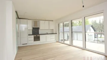 Heller offener Wohnbereich mit integrierter weißer Küche, Holzboden und Zugang zu einem großen Balkon.
