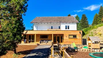 Modernes Gebäude mit Holzverkleidung und Satteldach, umgeben von Bäumen und einem Spielbereich.