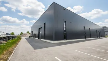 Moderne Gewerbehalle mit dunkler Fassade und großem Parkplatz unter blauem Himmel.