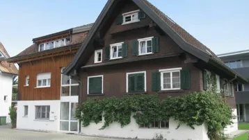 Traditionelles Haus mit dunkler Holzfassade, grünen Fensterläden und bewachsenem Sockelbereich.