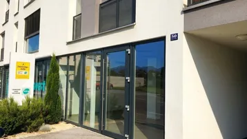 Moderne Gebäudefassade mit großen Glasfenstern und Eingangstüren, umgeben von Grünflächen.