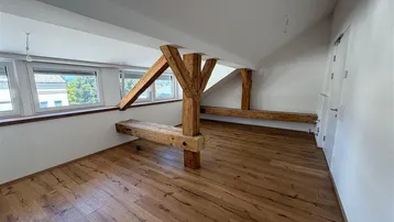 Geräumiger Dachbodenraum mit sichtbaren Holzbalken, Holzboden und mehreren Fenstern, die viel Tageslicht hereinlassen.