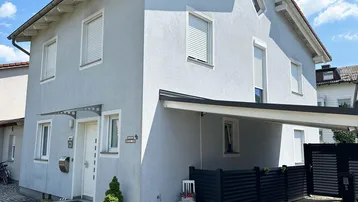 Graues Einfamilienhaus mit Rollläden und überdachtem Eingangsbereich unter blauem Himmel.