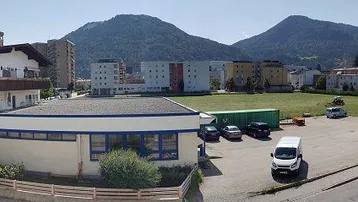 Panoramablick auf ein Gewerbegebäude mit Parkplatz, umgeben von Wohngebäuden und einer beeindruckenden Bergkulisse.