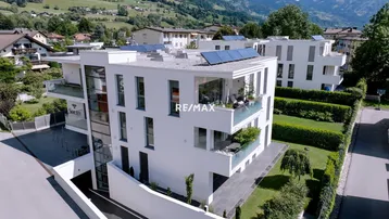 Moderne Wohnanlage mit weißer Fassade, Balkonen, Solarpaneelen und Bergblick in grüner Umgebung.