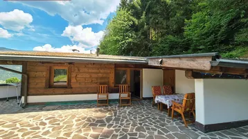 Große gepflasterte Terrasse mit Holzhütte und Sitzgelegenheiten, umgeben von grünen Bäumen und Bergen.
