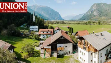 Luftaufnahme eines ländlichen Anwesens mit mehreren Gebäuden, umgeben von grünen Wiesen und Bergen unter blauem Himmel.